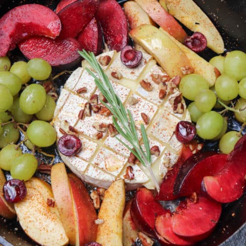 A cast iron skillet with a round wheel of brie cheese topped with rosemary and pecans, surrounded by sliced apples, plums, grapes, and cherries, all sprinkled with spices.