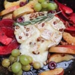 A cast iron skillet with baked brie topped with herbs and chopped nuts, surrounded by sliced apples, red plums, green grapes, and cherries. The fruit is arranged around the melted cheese.