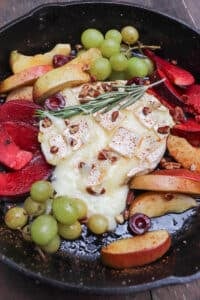 A cast iron skillet with baked brie topped with herbs and chopped nuts, surrounded by sliced apples, red plums, green grapes, and cherries. The fruit is arranged around the melted cheese.