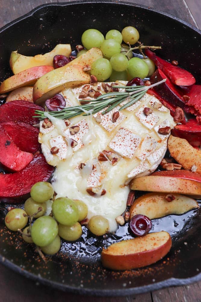 A cast iron skillet with baked brie topped with herbs and chopped nuts, surrounded by sliced apples, red plums, green grapes, and cherries. The fruit is arranged around the melted cheese.
