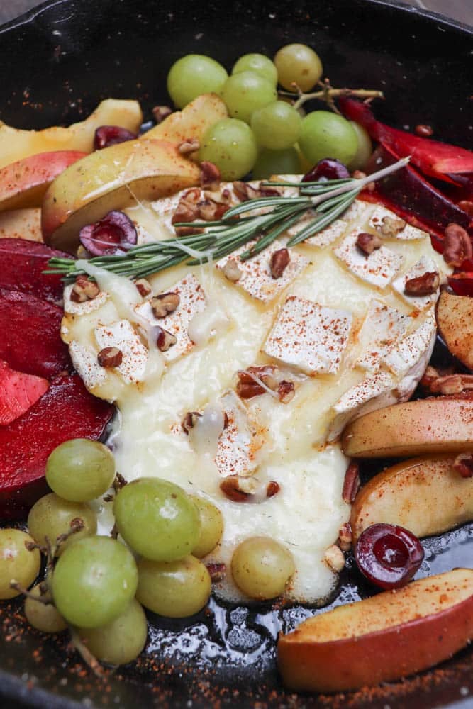 A round of baked brie topped with nuts and herbs, partially melted, surrounded by green grapes, plum slices, apple slices, and halved cherries in a skillet.