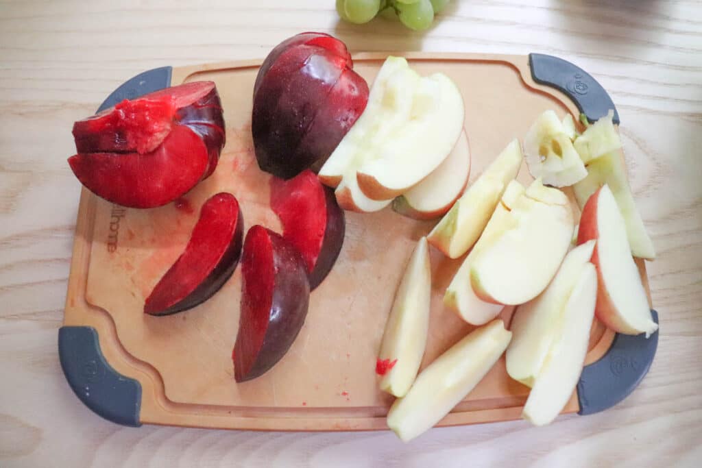 Sliced red plums and sliced apples are arranged on a wooden cutting board with gray corner grips; green grapes are visible in the background.