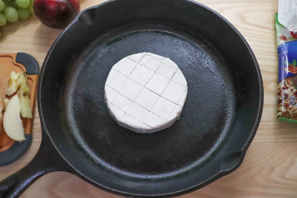 A round wheel of brie cheese with a crosshatch pattern cut into the top sits in the center of a cast iron skillet on a wooden surface. Sliced apples and grapes are visible nearby.