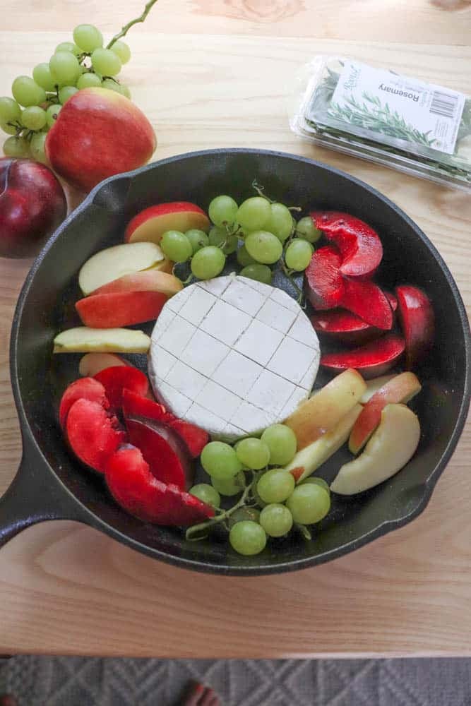 A cast iron skillet with a round wheel of cheese in the center, surrounded by sliced red plums, apple wedges, and green grapes. Fresh fruit and a rosemary package are on the wooden table nearby.