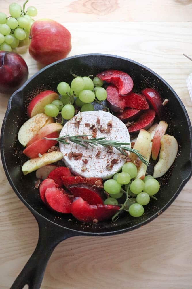 A cast iron skillet filled with sliced apples, plums, green grapes, and a round of brie cheese topped with herbs and chopped nuts, arranged on a wooden surface.