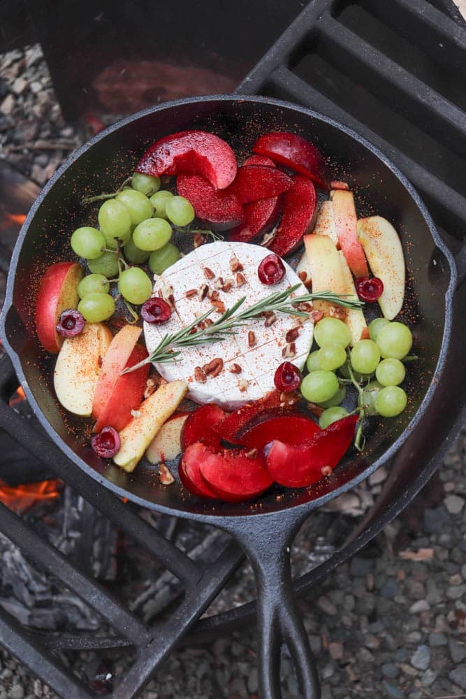 A cast-iron skillet over an open flame contains a wheel of cheese topped with herbs, surrounded by slices of plum, apple, grapes, cherries, and sprinkled spices.