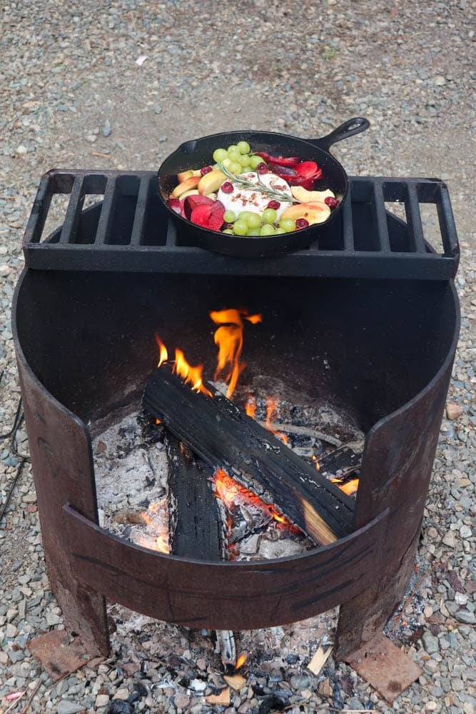 A cast iron skillet filled with fruit and what appears to be cheese sits on a grill over a campfire, with burning logs and ashes below, and gravel surrounding the fire pit.