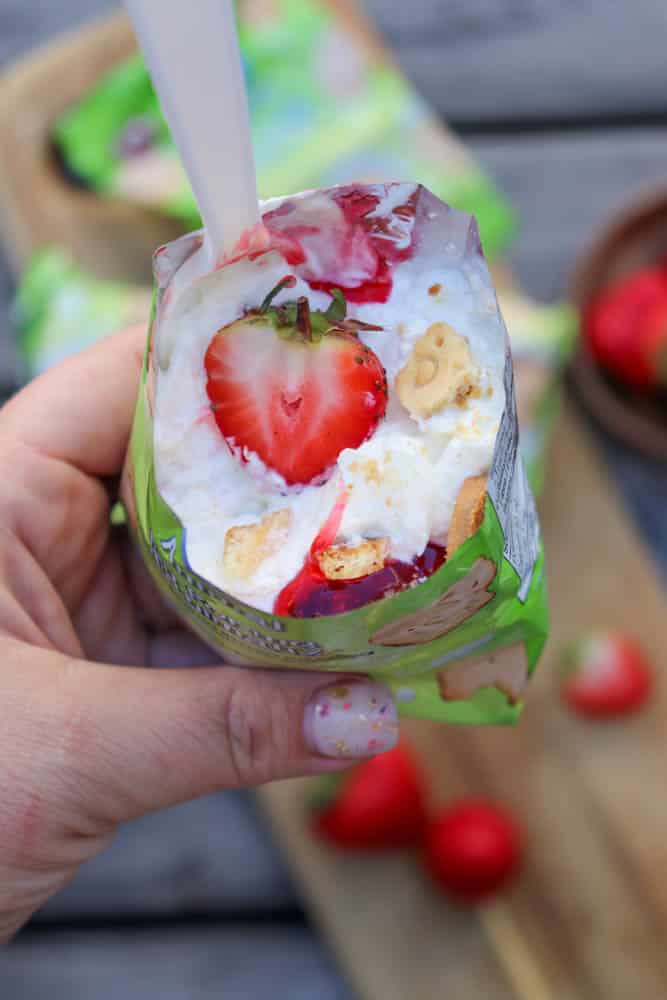 A hand holds an opened snack pouch filled with whipped cream, a whole strawberry, cookie pieces, and strawberry sauce; a plastic spoon is inside. More strawberries are on a wooden surface in the background.