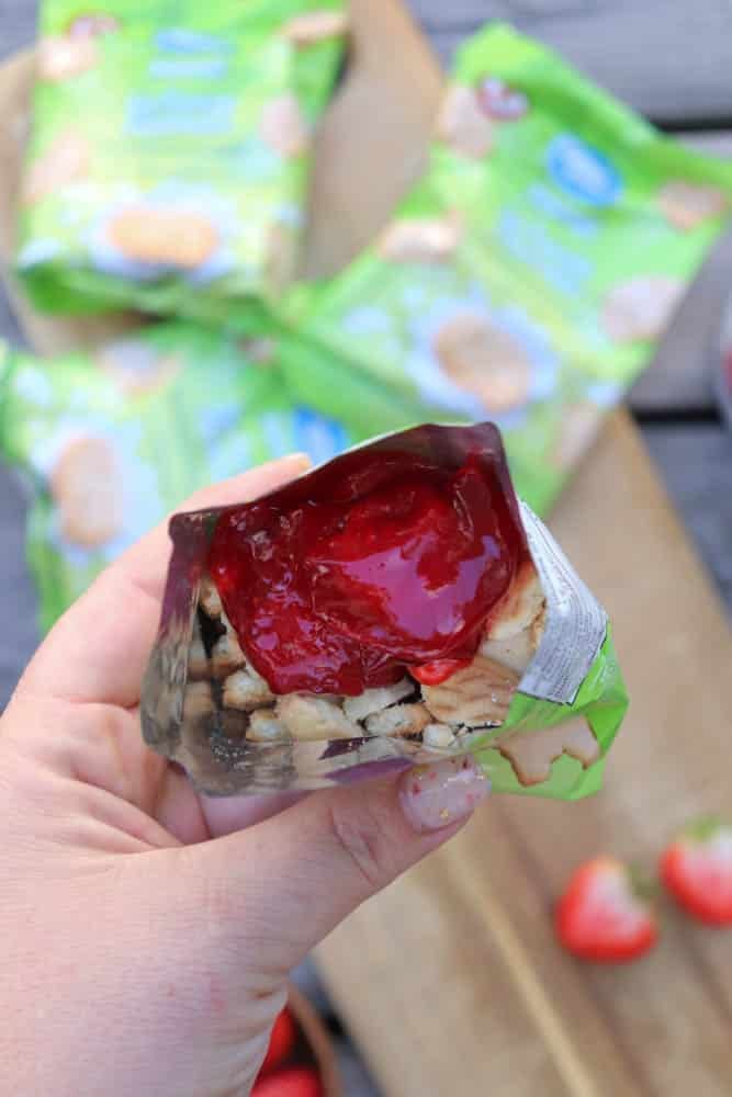 A hand holds an open snack pack filled with small biscuit pieces and topped with bright red strawberry jam, with more unopened packs and strawberries blurred in the background.