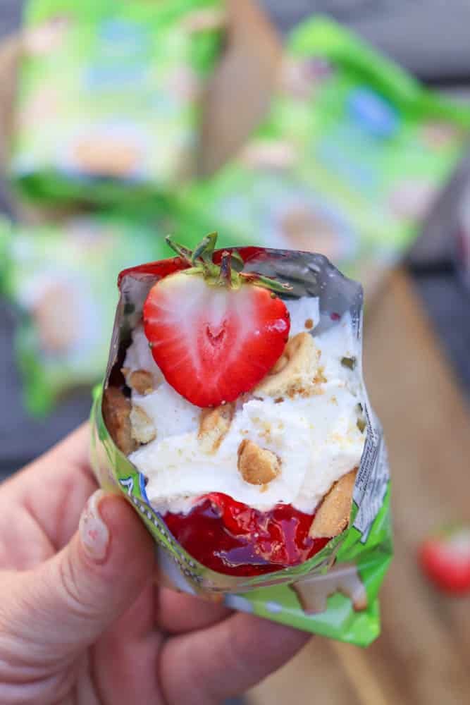 A hand holds an opened snack-sized bag filled with whipped cream, crumbled cookies, strawberry sauce, and topped with a halved fresh strawberry. Green snack packaging and blurred backgrounds are visible.