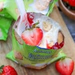 An opened green snack bag filled with vanilla pudding, animal-shaped cookies, and sliced strawberries, with a white plastic spoon inside. Fresh strawberries are placed nearby on a wooden surface.