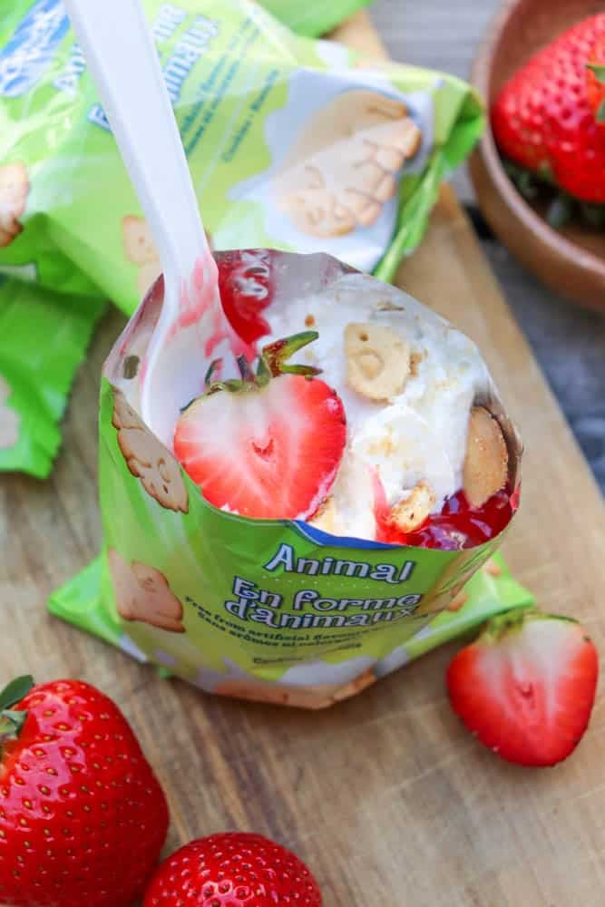 An opened green snack bag filled with vanilla pudding, animal-shaped cookies, and sliced strawberries, with a white plastic spoon inside. Fresh strawberries are placed nearby on a wooden surface.