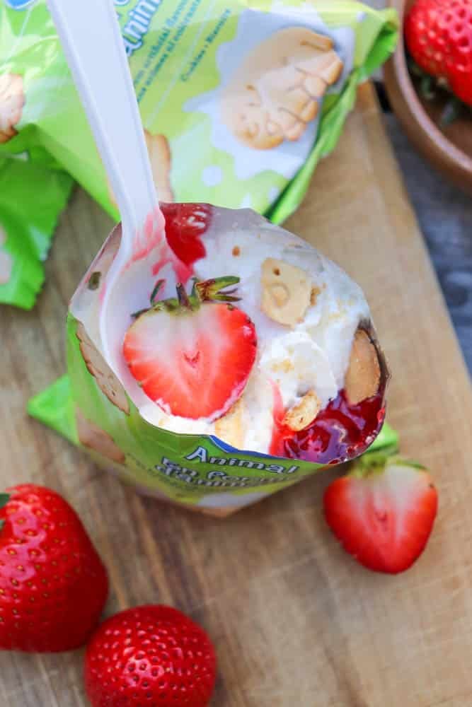 A partially opened snack pouch filled with vanilla ice cream, a whole strawberry, and small cookies sits on a wooden board next to fresh strawberries and a green snack wrapper. A white plastic spoon is in the pouch.