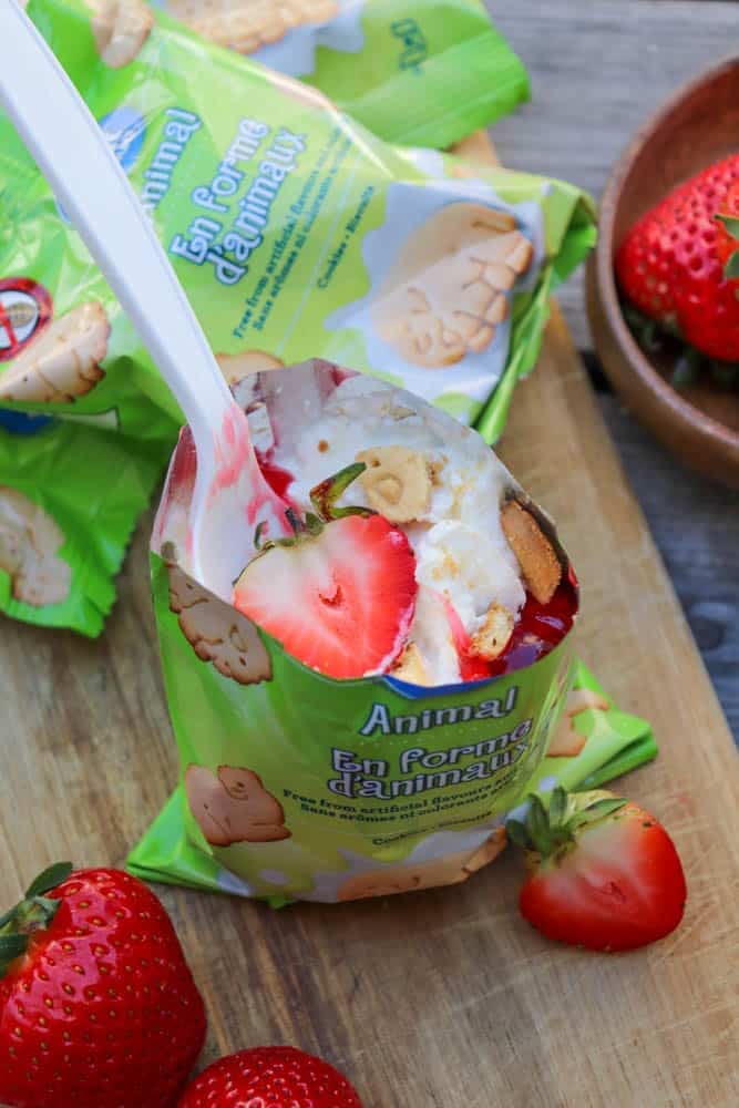 A cup filled with animal-shaped cookies, whipped cream, and strawberry slices sits on a wooden surface, surrounded by fresh strawberries and green animal cookie packages. A spoon is inside the cup.