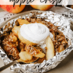 Apple crisp served in a foil packet, topped with whipped cream. The dessert sits on a beige plate with a spoon beside it. Fresh apples are in the background, and text reads Foil Packet Apple Crisp.
