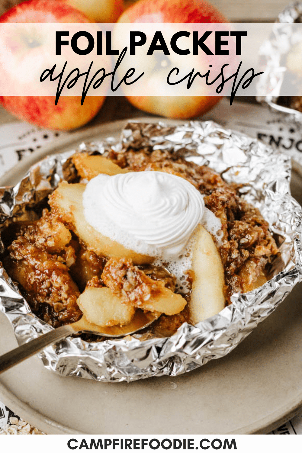 Apple crisp served in a foil packet, topped with whipped cream. The dessert sits on a beige plate with a spoon beside it. Fresh apples are in the background, and text reads Foil Packet Apple Crisp.