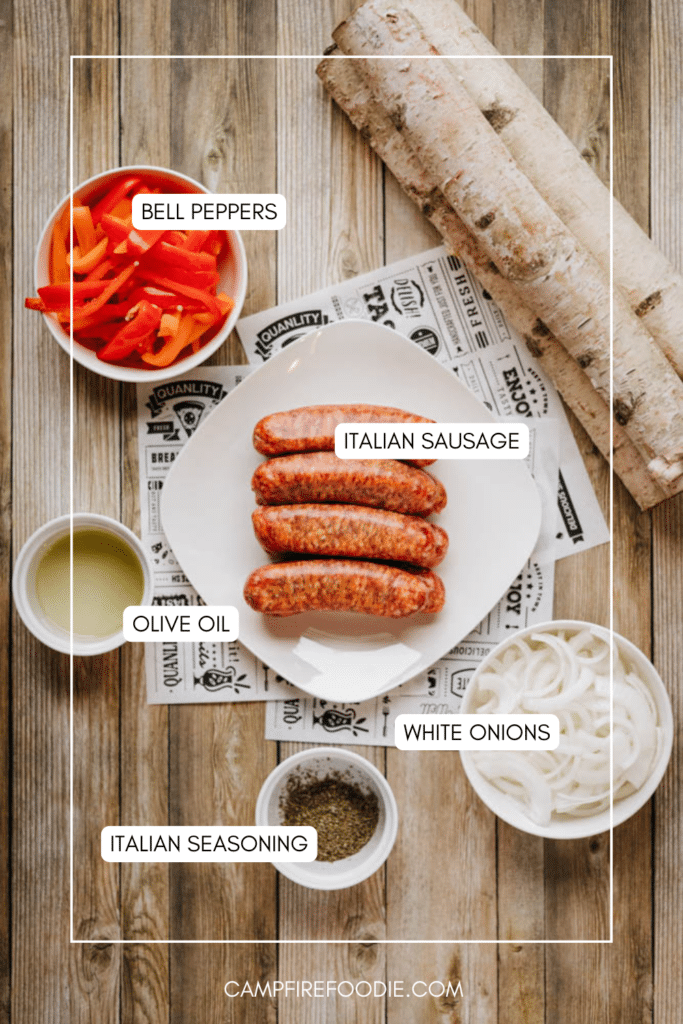 Top-down view of a plate with three Italian sausages, surrounded by bowls of sliced bell peppers, white onions, olive oil, Italian seasoning, and birch logs on a wooden surface. Labels identify each ingredient.