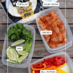 Labeled containers of sliced bell peppers, seasoned chicken, lime wedges with cilantro, a pack of keto tortillas, and a tub of sour cream arranged on a wooden surface.