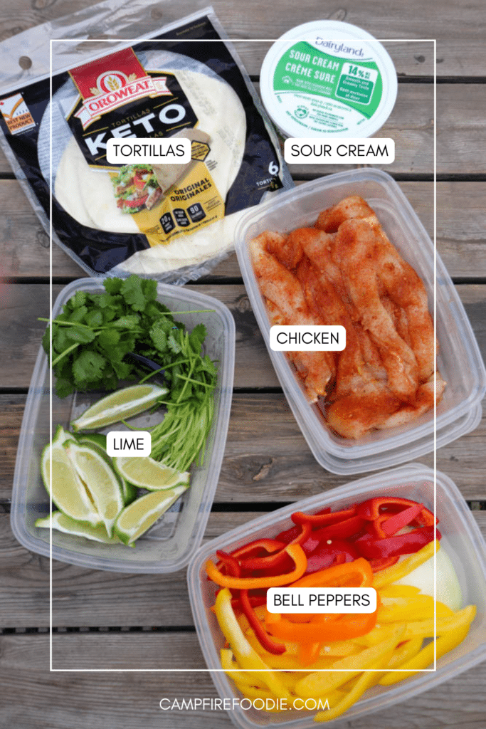 Labeled containers of sliced bell peppers, seasoned chicken, lime wedges with cilantro, a pack of keto tortillas, and a tub of sour cream arranged on a wooden surface.
