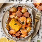 Foil packet filled with shrimp, sausage, corn on the cob, and baby potatoes on a plate with a fork. Slices of lemon and parsley garnish are beside it. Text reads “Foil Packet Shrimp Boil” and “campfirefoodie.com.”.