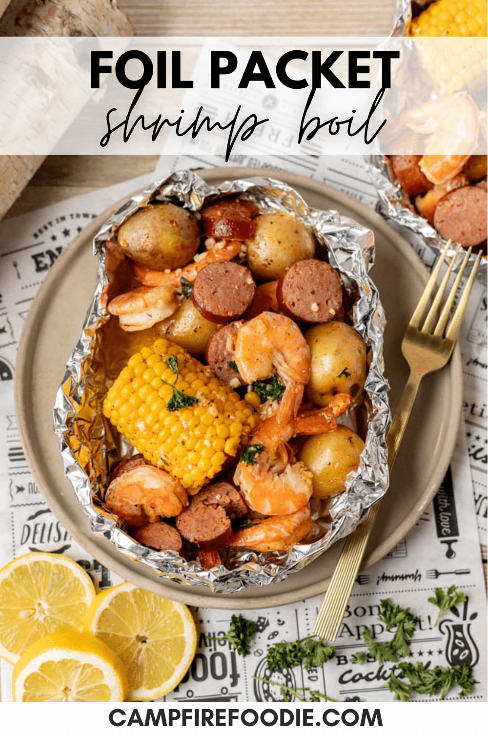 Foil packet filled with shrimp, sausage, corn on the cob, and baby potatoes on a plate with a fork. Slices of lemon and parsley garnish are beside it. Text reads “Foil Packet Shrimp Boil” and “campfirefoodie.com.”.