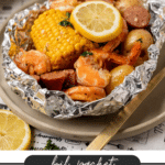 A foil packet on a plate filled with shrimp, corn on the cob, sausage slices, small potatoes, a lemon slice, and herbs. The text below reads: “foil packet shrimp boil - campfirefoodie.com.”.
