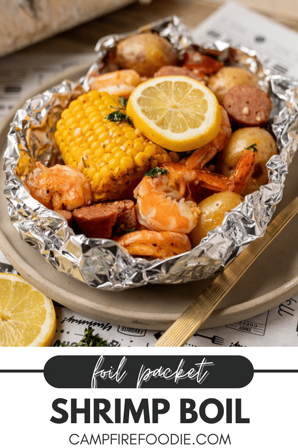 A foil packet on a plate filled with shrimp, corn on the cob, sausage slices, small potatoes, a lemon slice, and herbs. The text below reads: “foil packet shrimp boil - campfirefoodie.com.”.