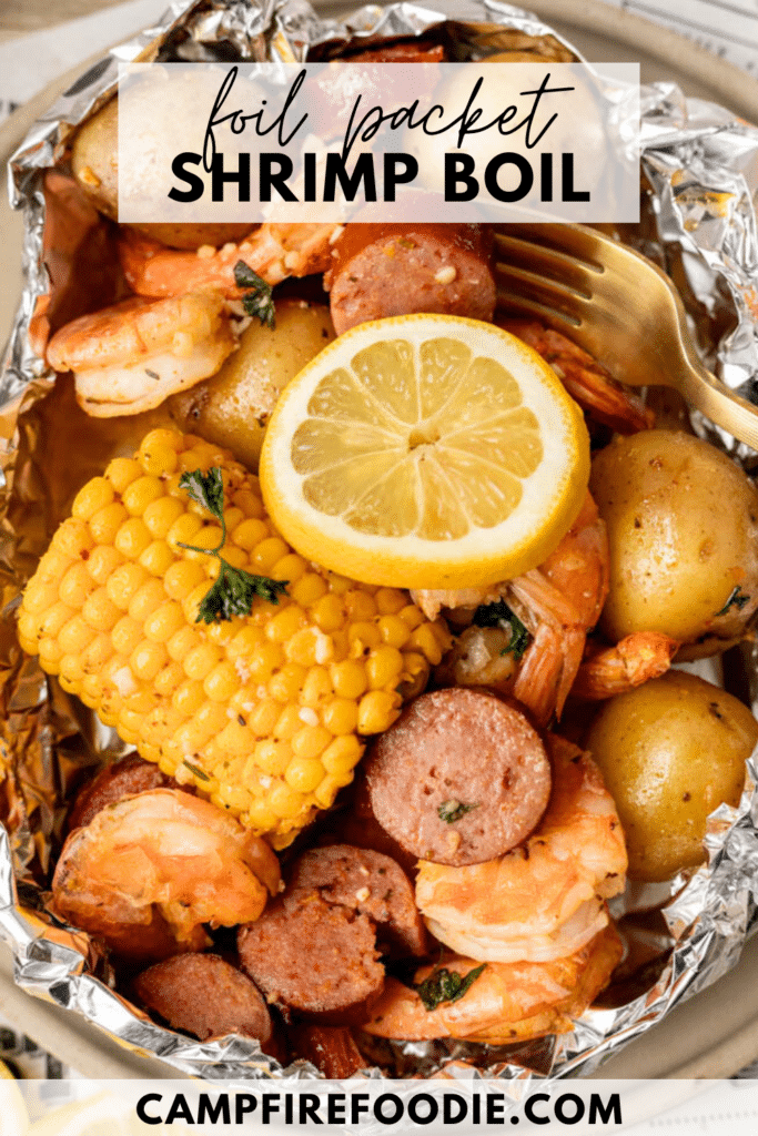 Foil packet filled with shrimp, sliced sausage, corn on the cob, potatoes, and a lemon slice, garnished with herbs and served with a fork. Text reads foil packet shrimp boil and campfirefoodie.com.