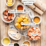 Top-down view of ingredients for a shrimp boil, including lemon slices, Mennonite sausage, mango chipotle seasoning, corn, potatoes, shrimp, butter, garlic, lemon, and fresh herbs, all labeled on a wooden table.