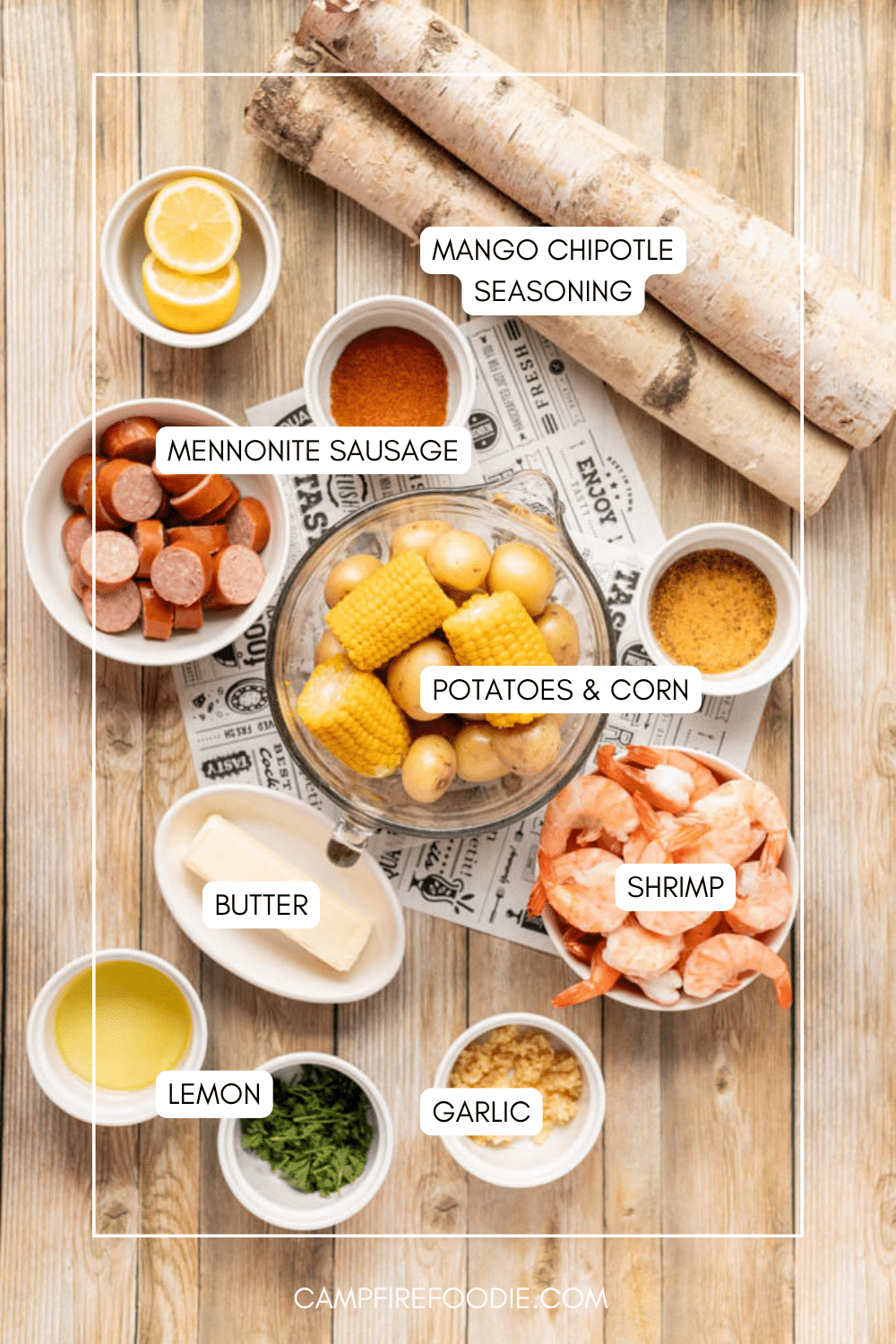 Top-down view of ingredients for a shrimp boil, including lemon slices, Mennonite sausage, mango chipotle seasoning, corn, potatoes, shrimp, butter, garlic, lemon, and fresh herbs, all labeled on a wooden table.