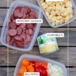 Three containers hold chopped hashbrowns, sliced Mennonite sausage, and diced bell pepper with onions, along with a can of corn niblets, all arranged on a wooden surface. Labels identify each ingredient.