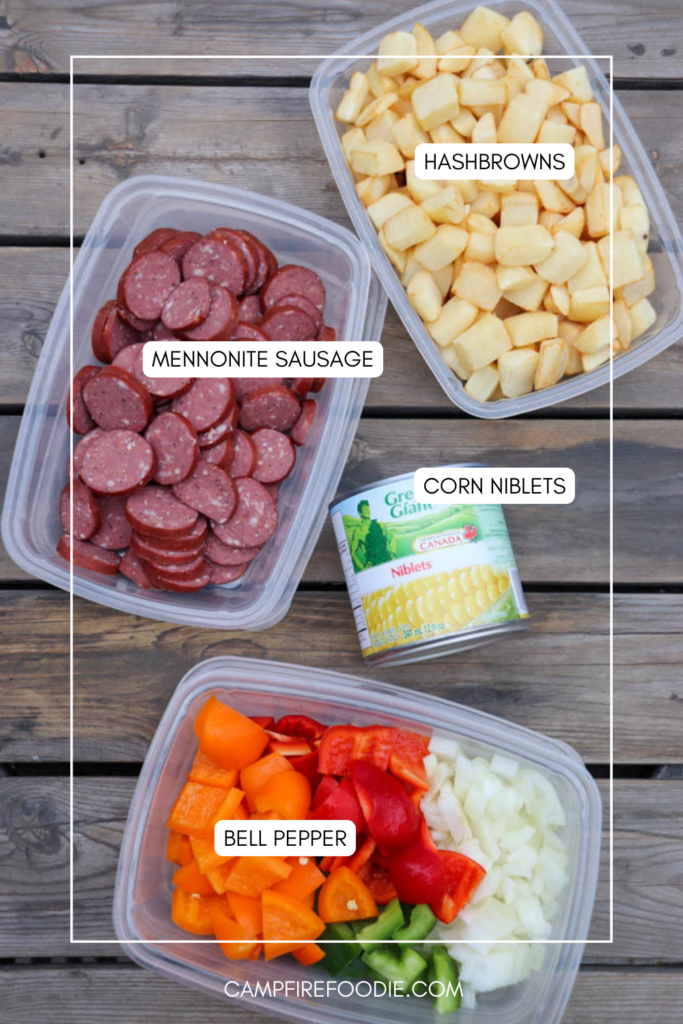 Three containers hold chopped hashbrowns, sliced Mennonite sausage, and diced bell pepper with onions, along with a can of corn niblets, all arranged on a wooden surface. Labels identify each ingredient.