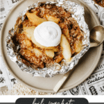 A foil packet filled with apple crisp topped with whipped cream sits on a plate with a spoon. The text below reads, foil packet APPLE CRISP campfirefoodie.com. Fresh apples are in the background.