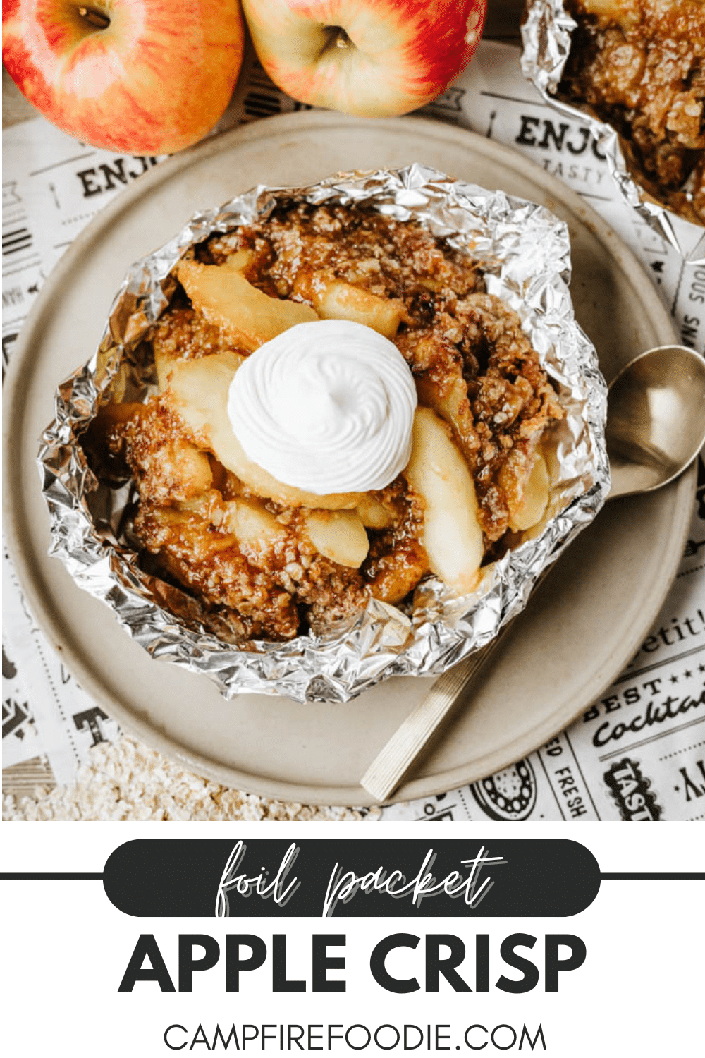 A foil packet filled with apple crisp topped with whipped cream sits on a plate with a spoon. The text below reads, foil packet APPLE CRISP campfirefoodie.com. Fresh apples are in the background.