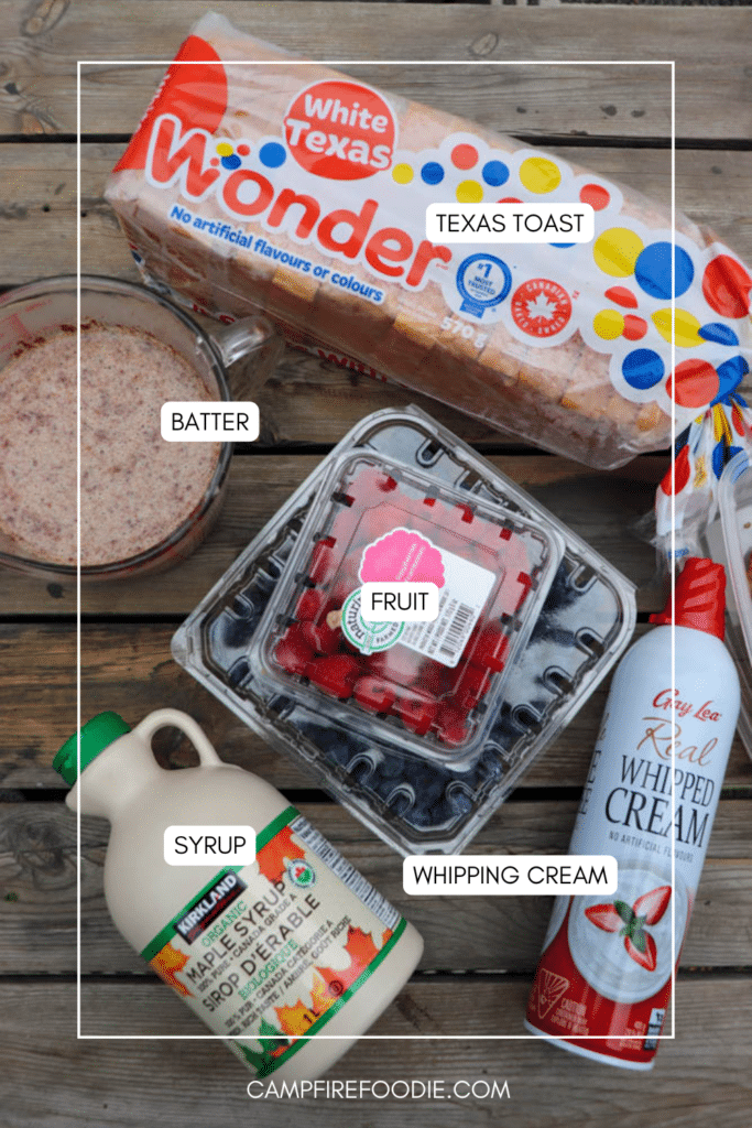A top-down view of ingredients on a wooden surface: a loaf of Texas toast bread, a clear container of berries, a can of whipped cream, a bottle of maple syrup, and a bowl of batter, each labeled.