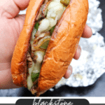 A hand holds a Philly cheesesteak sandwich filled with melted cheese, sautéed onions, green peppers, and steak in a toasted bun. Text below reads Blackstone Philly Cheesesteak, campfirefoodie.com.
