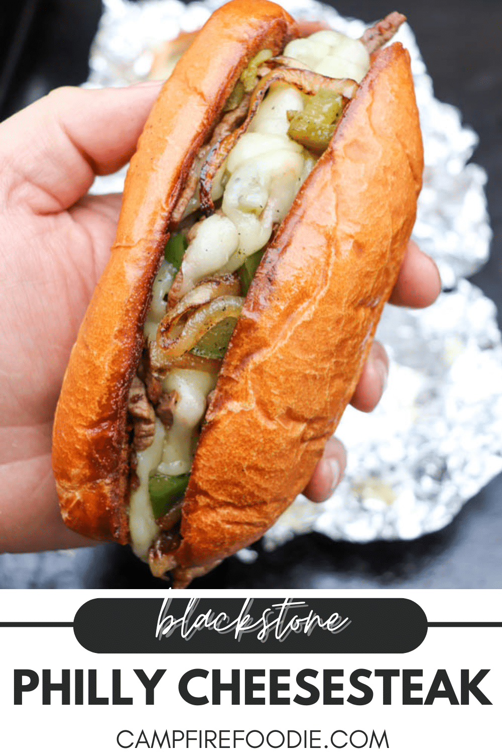 A hand holds a Philly cheesesteak sandwich filled with melted cheese, sautéed onions, green peppers, and steak in a toasted bun. Text below reads Blackstone Philly Cheesesteak, campfirefoodie.com.