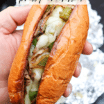 A hand holds a Philly cheesesteak sandwich with melted cheese, grilled onions, and green peppers in a toasted bun. Text on the image reads: Blackstone Philly Cheesesteak and campfirefoodie.com.