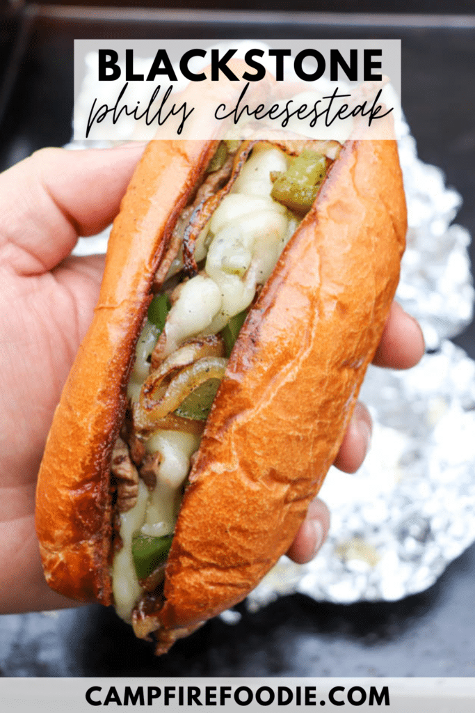A hand holds a Philly cheesesteak sandwich with melted cheese, grilled onions, and green peppers in a toasted bun. Text on the image reads: Blackstone Philly Cheesesteak and campfirefoodie.com.