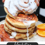 A hand lifts a frosted cinnamon roll from a stack of three on a plate, with strips of bacon and orange slices beside them. The text reads, Blackstone Cinnamon Rolls campfirefoodie.com.