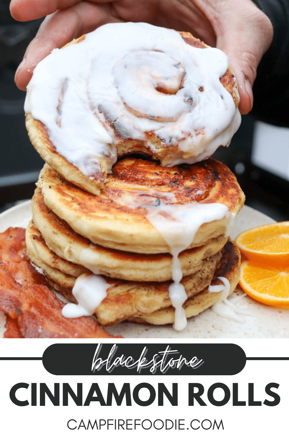 A hand lifts a frosted cinnamon roll from a stack of three on a plate, with strips of bacon and orange slices beside them. The text reads, Blackstone Cinnamon Rolls campfirefoodie.com.