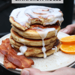 A hand holds a plate stacked with iced cinnamon roll pancakes, served with two strips of bacon and orange slices. The text reads “Blackstone Cinnamon Rolls” and “campfirefoodie.com.”.