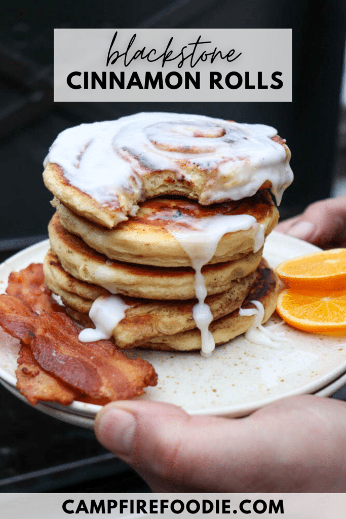 A hand holds a plate stacked with iced cinnamon roll pancakes, served with two strips of bacon and orange slices. The text reads “Blackstone Cinnamon Rolls” and “campfirefoodie.com.”.