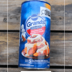 A can of Pillsbury Grands! Cinnabon Cinnamon Rolls is placed on a wooden surface. The label shows iced cinnamon rolls and the Pillsbury Doughboy mascot. Text: Grands Cinnamon Rolls.