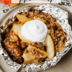A foil packet filled with apple crisp topped with whipped cream sits on a plate. A fork rests in the dessert, and fresh apples are visible in the background. Text reads Foil Packet Apple Crisp and campfirefoodie.com.