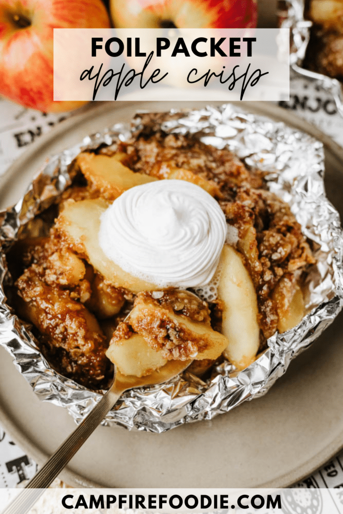 A foil packet filled with apple crisp topped with whipped cream sits on a plate. A fork rests in the dessert, and fresh apples are visible in the background. Text reads Foil Packet Apple Crisp and campfirefoodie.com.