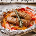 A foil packet filled with cooked sausages, red and yellow bell peppers, garnished with a sprig of rosemary, set on a plate. Text reads Foil Packet Sausage & Peppers and campfirefoodie.com.