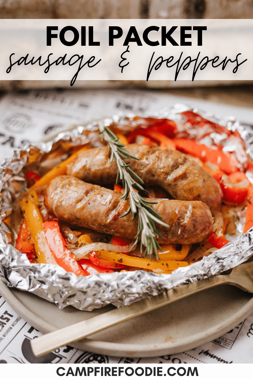 A foil packet filled with cooked sausages, red and yellow bell peppers, garnished with a sprig of rosemary, set on a plate. Text reads Foil Packet Sausage & Peppers and campfirefoodie.com.