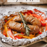 Two cooked sausages, sliced bell peppers, and onions are arranged in a foil packet, garnished with a sprig of rosemary. The foil sits on a plate with “Foil Packet Sausage & Peppers” text above and campfirefoodie.com below.