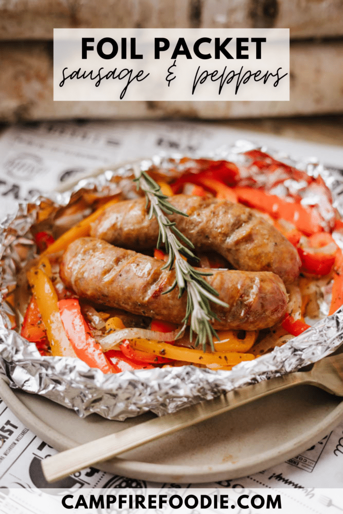 Two cooked sausages, sliced bell peppers, and onions are arranged in a foil packet, garnished with a sprig of rosemary. The foil sits on a plate with “Foil Packet Sausage & Peppers” text above and campfirefoodie.com below.